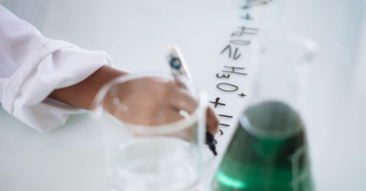 Body part of unrecognizable scientist in white uniform writing down formula after providing chemical research with fluid in flask during science lesson in university